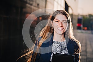 Successful beautiful young business woman is smiling on the background of buildings and holding a tablet computer.