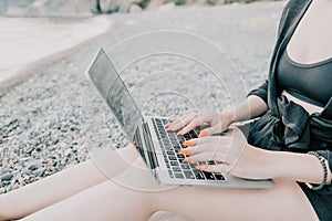 Successful business woman with bright red manicure typing on laptop keyboard outdoors on beach with sea view. Close up