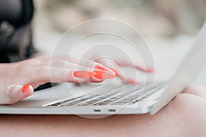 Successful business woman with bright red manicure typing on laptop keyboard outdoors on beach with sea view. Close up