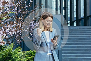 Successful business woman in a business suit outside the office reads good news from the phone, smiles and rejoices in victory, a