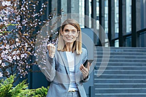 Successful business woman in a business suit outside the office reads good news from the phone, smiles and rejoices in victory, a