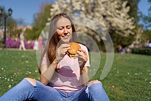 Successful business woman eating fast food burger cheesburger enjoys her leisure free time in a park with blossoming