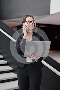 successful business woman standing with laptop in hands, working, talking on phone with colleagues