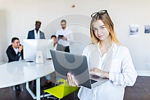 Successful business woman standing with laptop with her staff in background at modern bright office