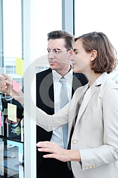 Successful businessman and businesswoman writing good ideas on sticky notes pasted on glass wall in modern office meeting