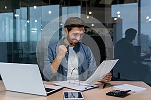 Successful businessman in casual shirt doing paperwork, boss with beard and glasses sitting at desk at workplace using