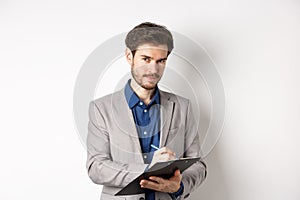 Successful businessman in grey suit taking notes, working in office clothing, holding clipboard and looking at camera