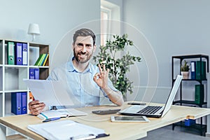 Successful businessman looks at the camera and smiles, performs reconciliation of business reports, shows the camera gesture ok