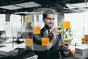 Successful businessman rewrites a task on a glass board with colored stickers, from phone and tablet