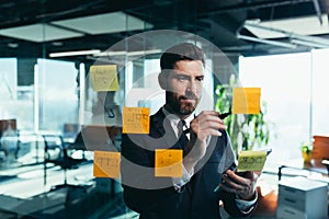 Successful businessman rewrites a task on a glass board with colored stickers, from phone and tablet