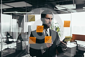 Successful businessman rewrites a task on a glass board with colored stickers, from phone and tablet