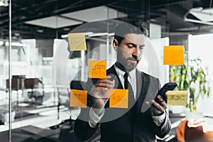 Successful businessman rewrites a task on a glass board with colored stickers, from phone and tablet