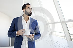 Successful businessman with a suit drinks coffee in the morning and looks out the window, an employee is standing in a white