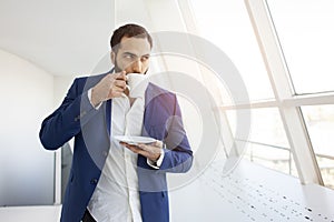 Successful businessman with a suit drinks coffee in the morning and looks out the window, an employee is standing in a white