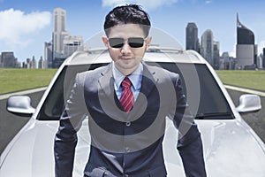 Successful businessman wearing sunglasses in front of luxury car with cityscape background