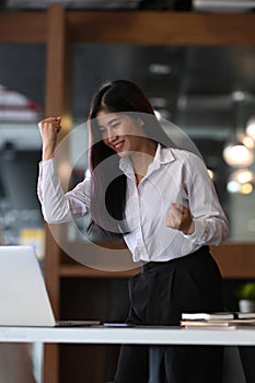 Successful businesswoman looking at laptop and raising her arm up to celebrating her success.