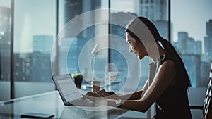 Successful Businesswoman in Stylish Dress Sitting at a Desk in Modern Office, Using Laptop Computer