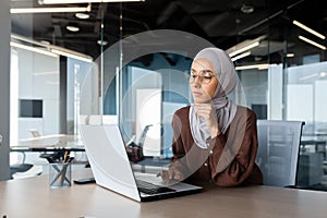 Successful businesswoman thinking at workplace, muslim woman in hijab using laptop at work, serious arab woman sitting