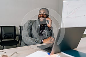 Successful cheerful African businessman in suit talking on smartphone sitting at office desk with laptop. Happy