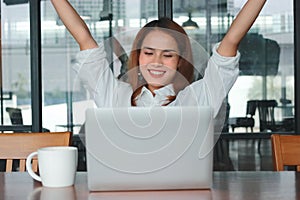 Successful cheerful young Asian businesswoman with laptop raising hands in office. Thinking and thoughtful business concept.