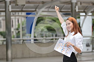 Successful cheerful young Asian businesswoman raising hands outside office. Thinking and thoughtful business concept