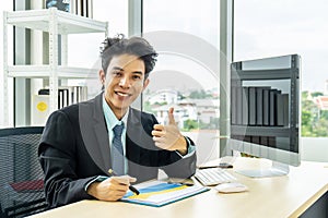 Successful confident asian businessman looking at camera  showing thumbs up or like while using laptop in office