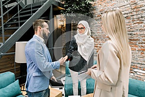 Successful confident Muslim business woman shaking hands with her two caucasian partners, blond woman and bearded man