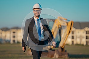 Successful construction business owner. Construction worker in suit and helmet near excavator. Confident construction