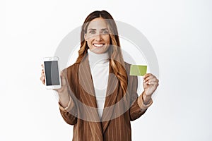 Successful corporate woman showing smartphone empty screen and credit card, app interface with order function, white