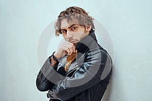 Daring and cool male model with curly hair posing in leather coat in a bright studio and leaning on the wall