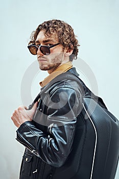 Portrait of a handsome guy with curly hair posing in the bright studio wearing sunglasses, leather coat and a backpack