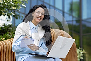 Successful diverse entrepreneur sitting on wooden bench and making notes in daybook while holding computer on laps