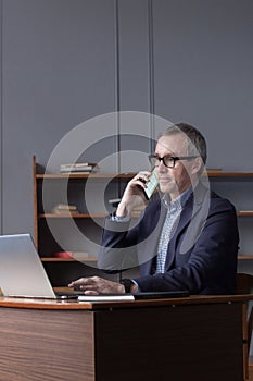 Successful elderly businessman in glasses typing on laptop and talking on mobile phone on office wall background