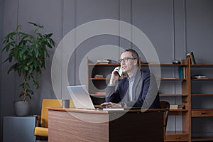 Successful elderly gray haired businessman in glasses working on laptop, talking on mobile phone on office wall background