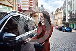 A Successful european young woman with long hair in a red coat talks on the phone and gets into her car