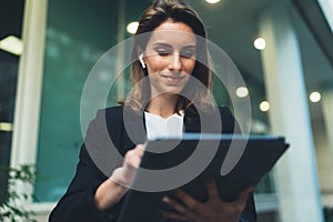 Successful female banker using tablet and wireless earphones outdoors near his office background lights, portrait young woman