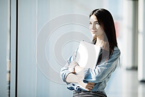 Successful female office worker with laptop is standing in skyscraper interior against big window with city view on background.
