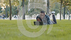 Successful freelancer business man teacher student african american guy lying on green grass lawn park having online