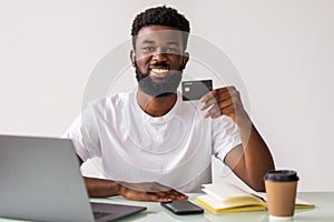 Successful freelancer receive payment. Happy African American man holding credit card, using laptop computer for online shopping