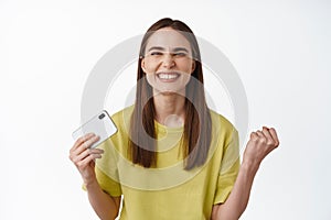 Successful girl looks satisfied, holds smartphone and fist pump, smiling pleased, celebrating victory, winning on phone