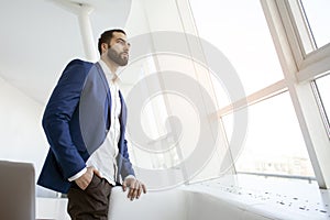 Successful handsome businessman in a blue suit stands against the background of a white office and looks out the window, portrait