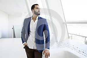 Successful handsome businessman in a blue suit stands against the background of a white office and looks out the window, portrait