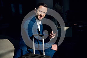 Successful handsome confident happy young businessman entrepreneur with mobile phone and suitcase smiles looking at camera sitting