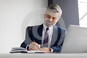 Successful handsome middle aged businessman writing in notepad while working in office