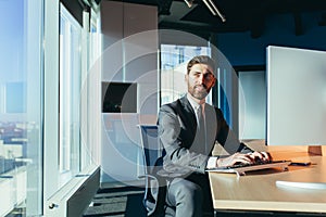 Successful and happy bearded man, working on a computer in a modern office by the window, businessman looking back and smiling