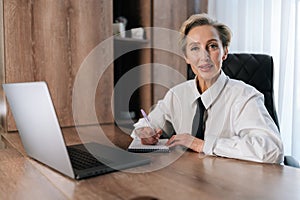 Successful and happy middle-aged business woman looking at camera sitting inside office working with documents. Portrait