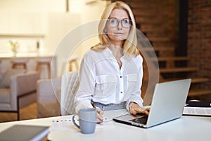 Successful mature businesswoman in glasses looking at camera, using laptop while doing some paperwork at the office