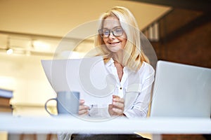 Successful mature businesswoman in glasses smiling, using laptop and doing some paperwork at the office