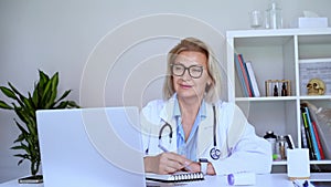 Successful mature caucasian senior female doctor in white coat using laptop for telemedicine with patients at hospital