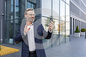 Successful mature man in business suit using app on phone for video call, senior businessman outside office building